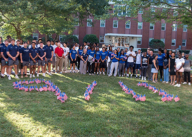 Students, faculty and staff gather in academic quad behind small American flags in the ground in the shape of 9/11