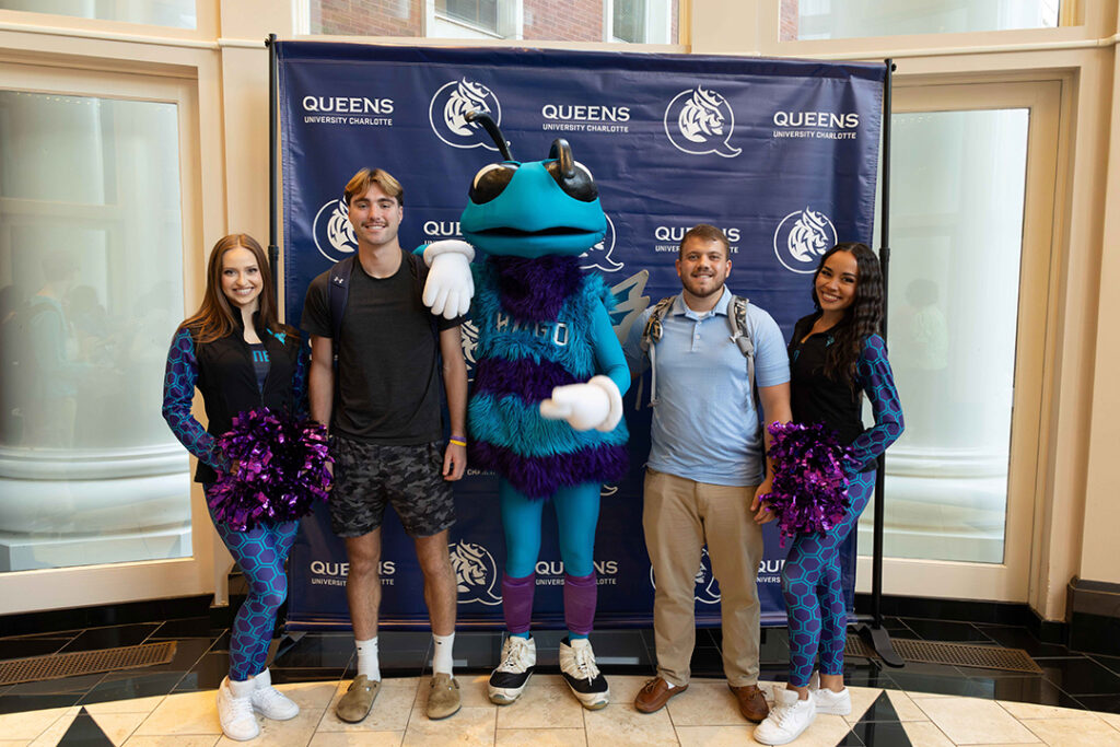 Two female dancers in teal and purple leotards flock male students with blue and teal bee Charlotte Hornets Mascot, mascot, Hugo.